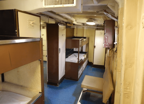 Interior of a ship's crew quarters, featuring bunk beds, lockers, and a small seating area.