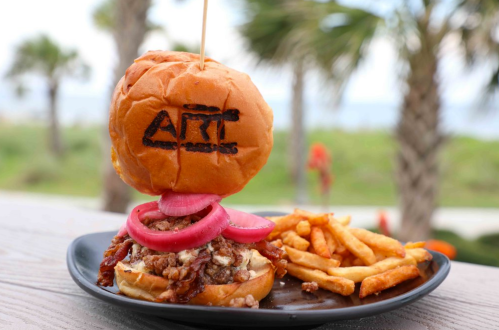 A gourmet burger with pickled onions and fries, featuring "ART" branded on the bun, set against a palm tree backdrop.
