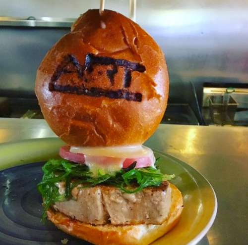 A burger with a toasted bun featuring "ART" branding, layered with greens, tomato, and a thick slice of meat.