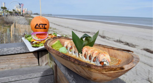A wooden platter with sushi and a salad, set against a beach backdrop with a beachside attraction in the distance.