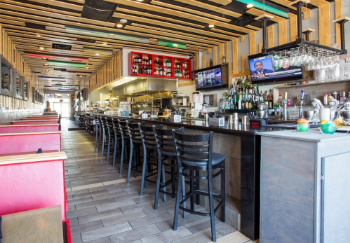 Interior of a modern restaurant featuring a long bar, seating, and large windows with TVs displaying news.
