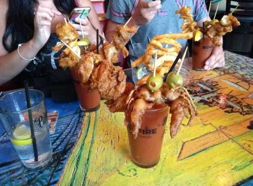 A colorful table with two elaborate cocktails topped with fried shrimp, olives, and curly fries, surrounded by people.