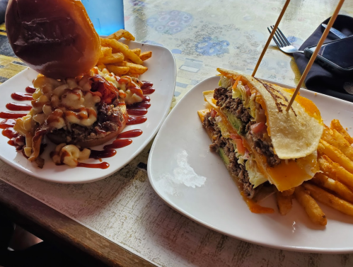 A plate with a stacked burger topped with mac and cheese and fries, alongside a loaded sandwich with fries.