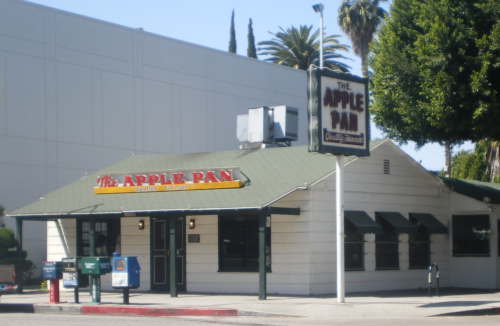 A small, charming restaurant called The Apple Pan, featuring a green roof and outdoor seating, surrounded by palm trees.