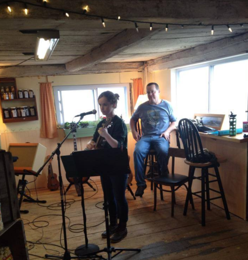 A woman plays guitar while singing on stage, with a man sitting on a stool nearby in a cozy, rustic setting.