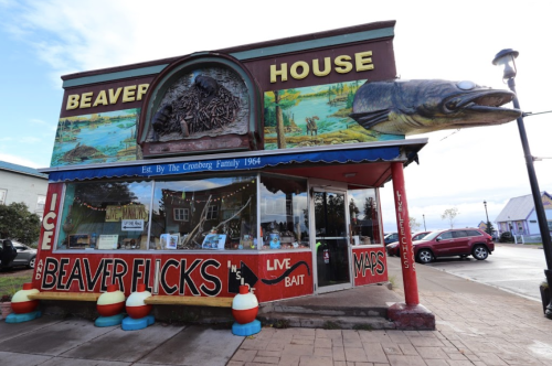 Colorful storefront of Beaver House, featuring a large fish sculpture and signs for ice, bait, and maps.