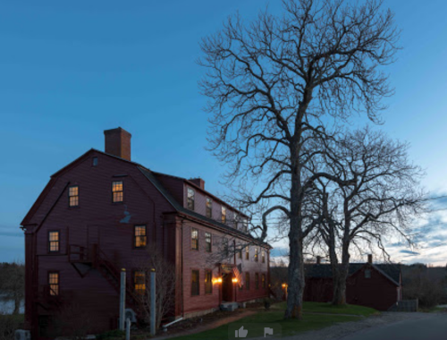 A red historic building at dusk, with large trees nearby and warm lights glowing from the windows.