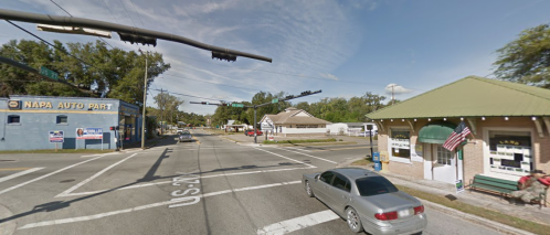 A street view showing a traffic intersection with a NAPA Auto Parts store and a small building with an American flag.