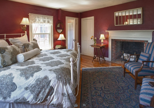 Cozy bedroom with a white bed, patterned bedding, a fireplace, and a vintage armchair against a deep red wall.