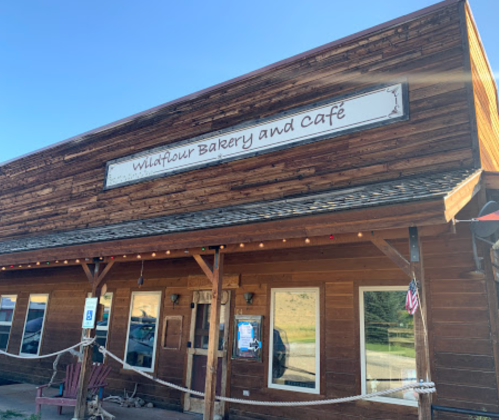 A rustic wooden building with a sign reading "Wildflour Bakery and Cafe," featuring a porch and decorative lights.