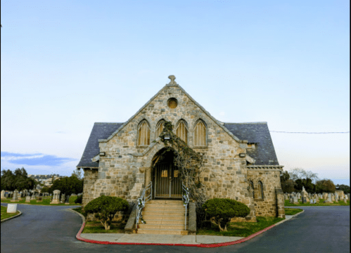 A stone chapel with arched windows, surrounded by neatly trimmed bushes, set against a clear sky.
