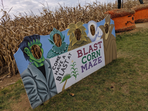 A colorful sign for a corn maze featuring cut-out holes for photos, with cartoon characters and the text "Blast Corn Maze."