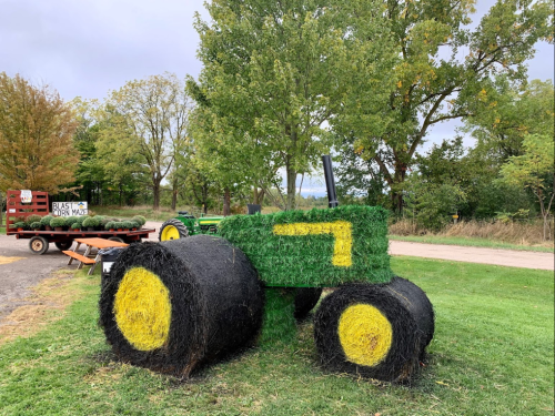 A large tractor sculpture made of hay bales, with green and yellow colors, set in a grassy area with trees in the background.