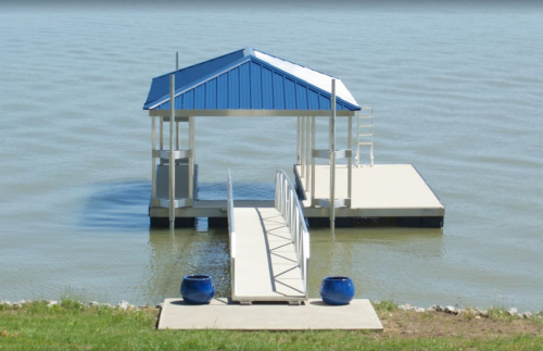 A small dock with a blue-roofed gazebo over calm water, featuring a walkway and two blue planters on the grass.