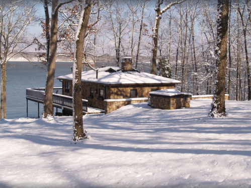 A cozy stone cabin surrounded by snow-covered trees, overlooking a serene lake in winter.