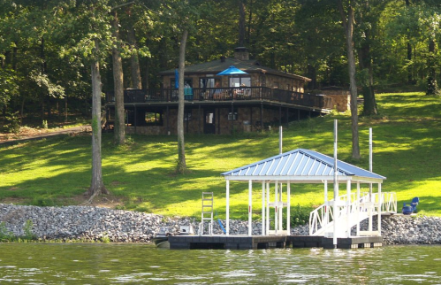 A lakeside view of a stone house surrounded by trees, with a dock extending into the water.