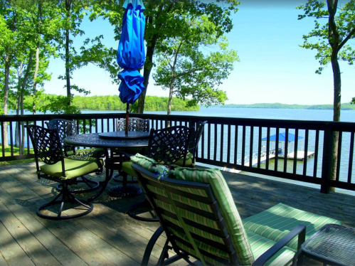 A lakeside deck with a table and chairs, surrounded by trees and a clear blue sky, overlooking the water.