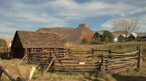 A rustic wooden cabin with a fenced area, set against a backdrop of a rocky mountain and blue sky.