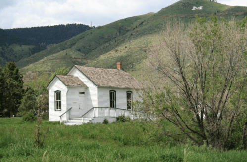 A small white house with a porch, surrounded by green grass and trees, set against a mountainous backdrop.