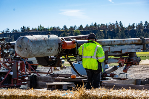A worker in a bright yellow jacket operates a large piece of machinery in a field under a clear blue sky.