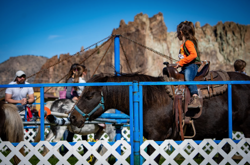 A child rides a pony at a fair, with mountains in the background and people enjoying the event nearby.