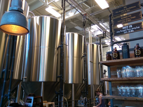 Interior of a brewery featuring large stainless steel fermentation tanks and a wooden bar with beer taps and bottles.