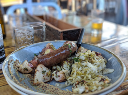 A plate with sausage, roasted potatoes, and sautéed cabbage on a wooden table, with drinks in the background.