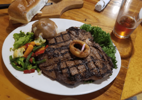 A grilled steak with a candle, served with mixed vegetables, a baked potato, and a roll, alongside a glass of beer.