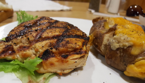 Grilled chicken breast on lettuce next to a baked potato topped with melted cheese and seasoning.