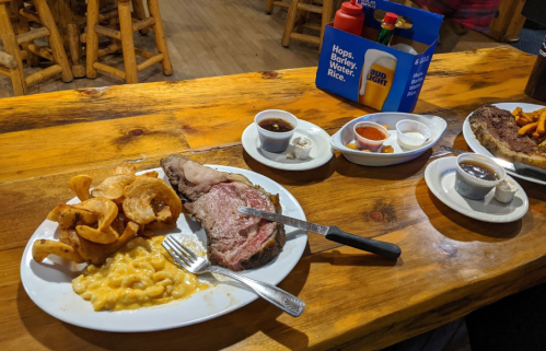 A plate with sliced prime rib, macaroni and cheese, and potato chips, alongside dipping sauces and drinks on a wooden table.
