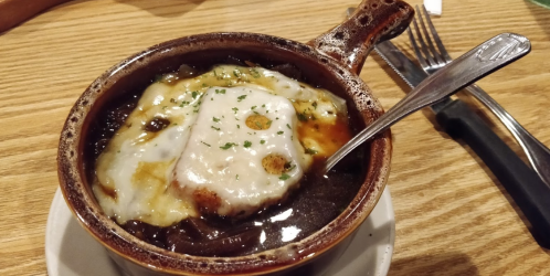 A bowl of French onion soup topped with melted cheese and herbs, served with a spoon on a wooden table.