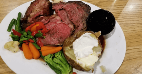A plate with sliced prime rib, a baked potato with sour cream, and a variety of steamed vegetables.