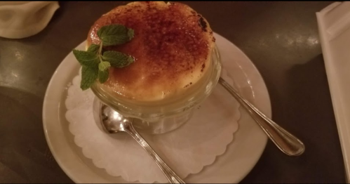A creamy dessert in a glass jar, topped with caramelized sugar and a mint sprig, served on a white plate with spoons.
