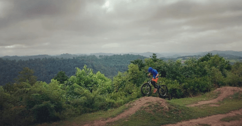 A mountain biker in blue gear rides over a dirt mound, surrounded by lush green hills under a cloudy sky.