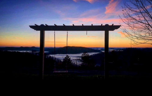 A silhouette of a swing set against a colorful sunset over misty hills and valleys.