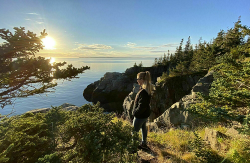 A person stands on a rocky ledge by the water, surrounded by trees, as the sun sets over the calm sea.