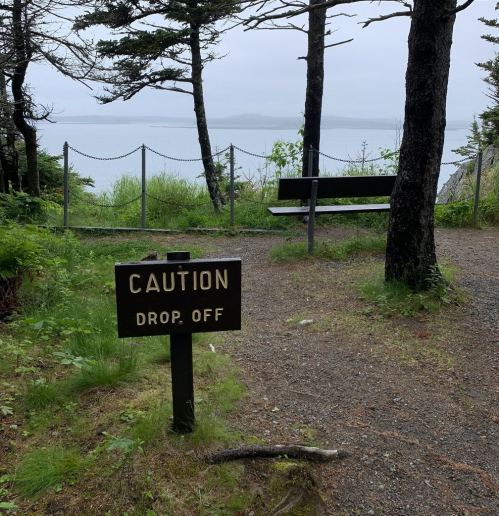 A sign reading "Caution: Drop Off" near a bench overlooking a foggy body of water, surrounded by trees.