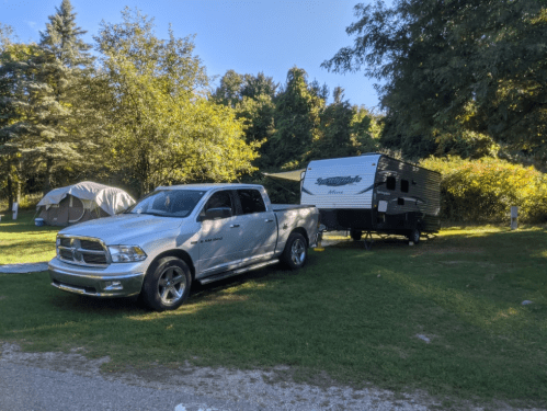 A silver pickup truck towing a black travel trailer, parked in a grassy campsite surrounded by trees.