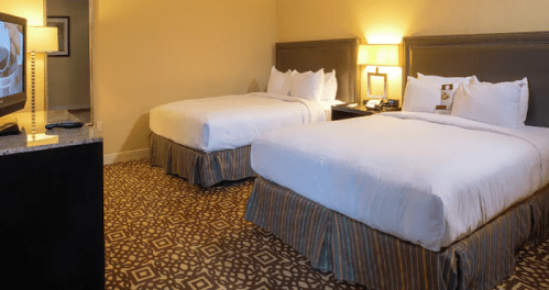 A hotel room featuring two queen beds, a TV, and a nightstand, with warm lighting and patterned carpet.