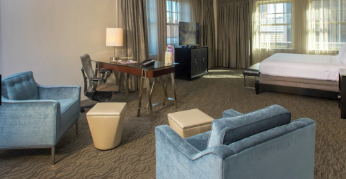 A spacious hotel room featuring a bed, desk, TV, and two blue chairs with a small ottoman. Natural light from large windows.