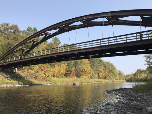 A metal arch bridge spans a calm river, surrounded by trees with autumn foliage in vibrant colors.
