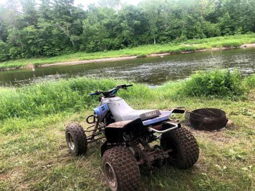 An ATV parked by a river, surrounded by lush greenery and a fire pit nearby.
