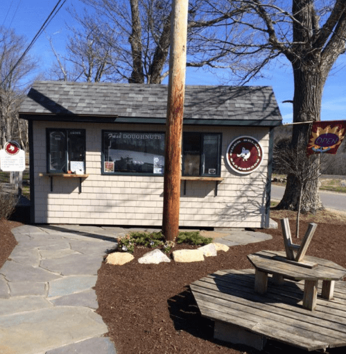 A small, charming doughnut stand with a wooden exterior, surrounded by trees and a flag indicating it's open.