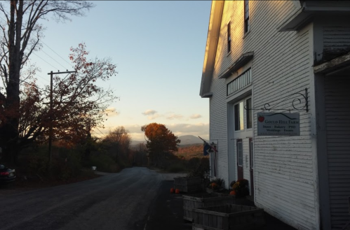 A rustic farm building at sunset, surrounded by trees and mountains, with a quiet road leading away.