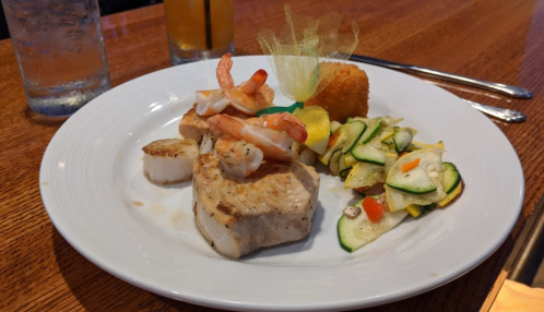 A plate of grilled fish topped with shrimp, scallops, and a side of sautéed vegetables, served with a croquette.