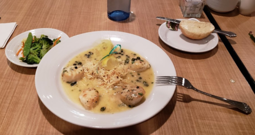 A plate of seafood dumplings in a creamy sauce, served with a side of broccoli and a bread roll on a wooden table.
