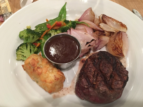 A plate featuring a grilled steak, roasted onions, broccoli, snap peas, and a side of sauce with a potato cake.