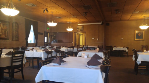 Interior of a restaurant with empty tables, white tablecloths, and warm lighting from chandeliers.
