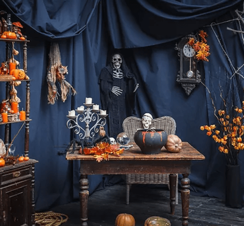 A spooky Halloween scene with a table, skeleton, pumpkins, candles, and autumn decorations against a dark backdrop.