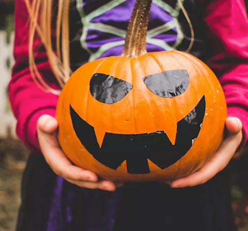 A child holds a carved pumpkin with a spooky face, featuring sharp teeth and angry eyes, dressed in a Halloween costume.